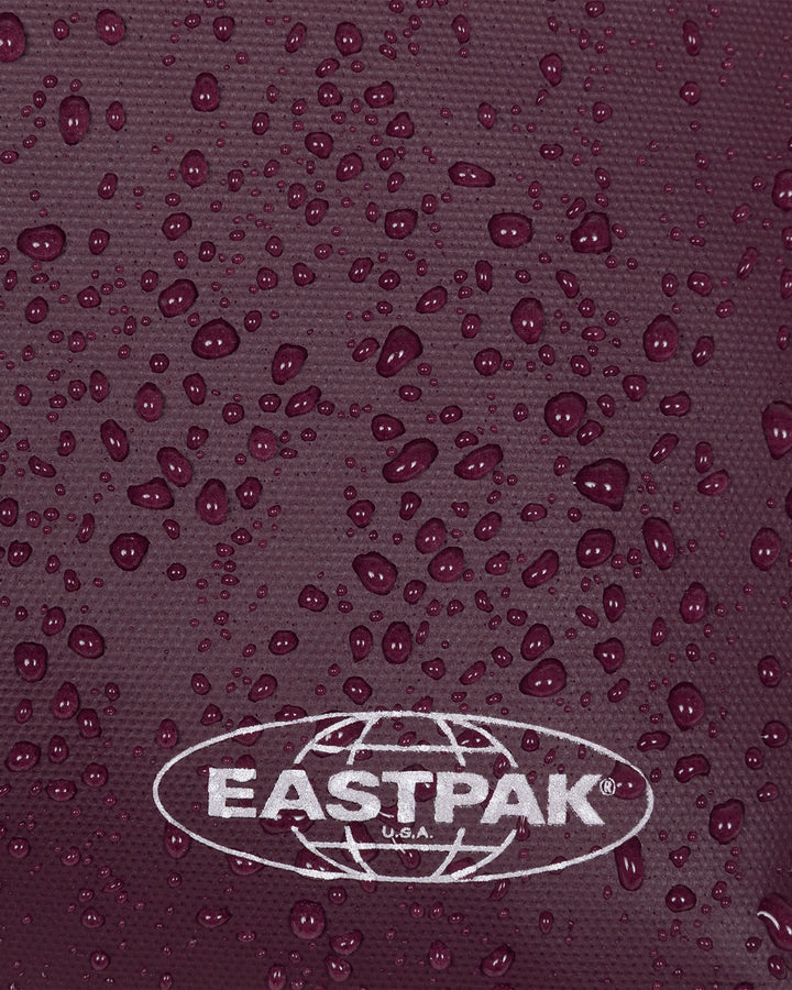 Close-up of a dark plum textured fabric covered with water droplets, featuring a white Eastpak logo at the bottom.