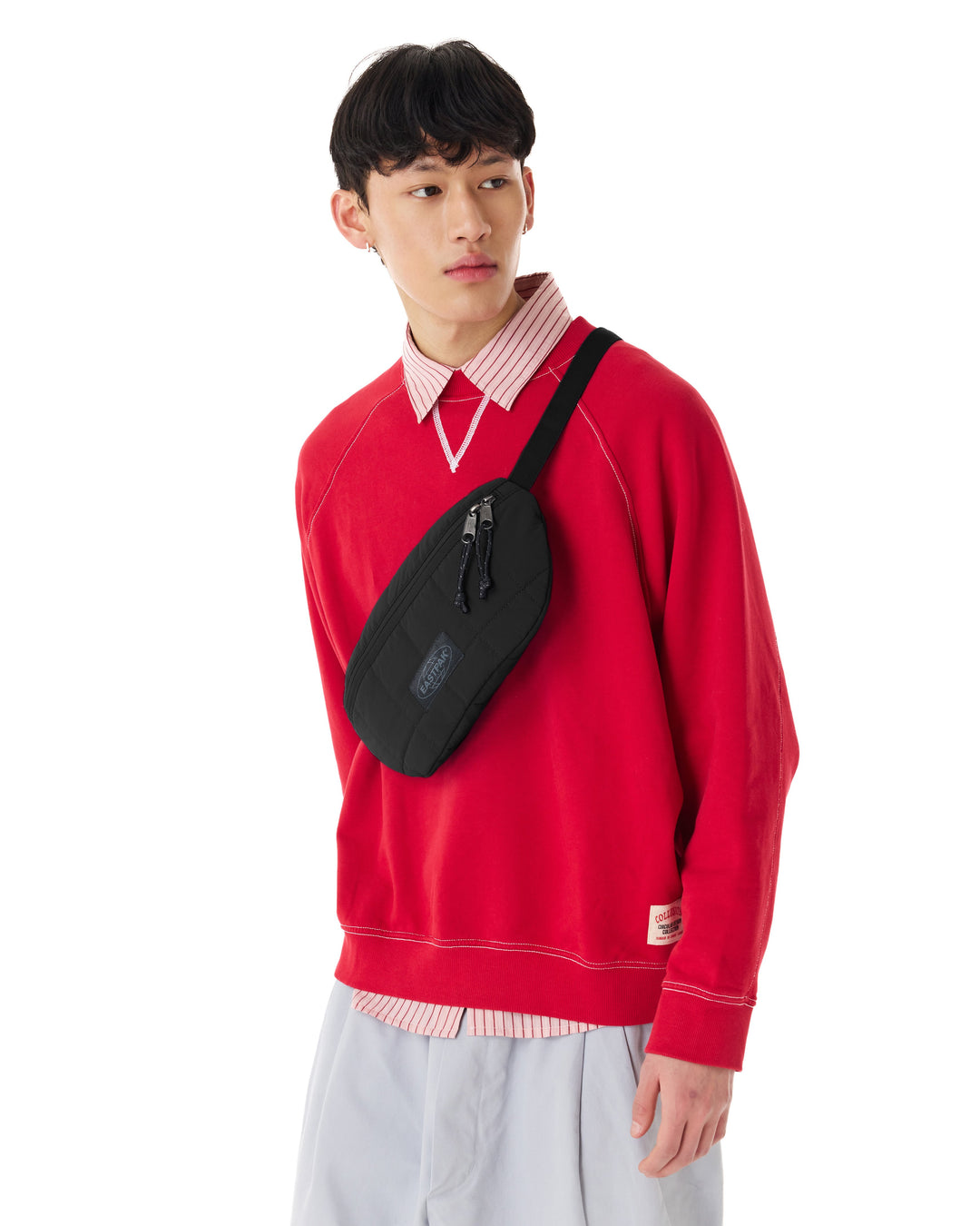 A model wearing a bright red crewneck sweatshirt, a pink and white striped collared shirt, and light gray pants, carrying a black crossbody sling bag across his chest against a plain white background.