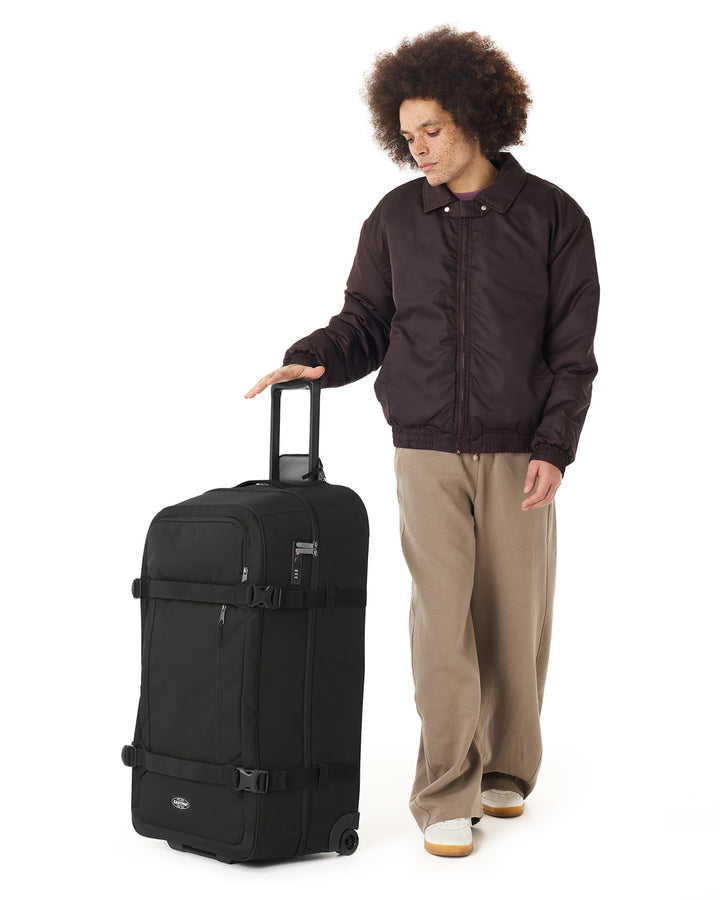 Person in a dark jacket and beige pants standing next to a black suitcase on a white background