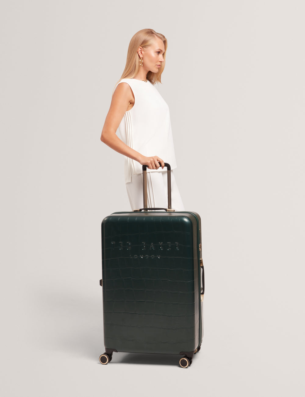 Model walking with a dark green suitcase with Ted Baker branding on a light grey background