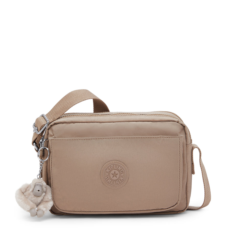 A taupe beige Kipling crossbody bag with a shoulder strap, a small monkey keychain charm, and a circular embossed Kipling logo on the front.