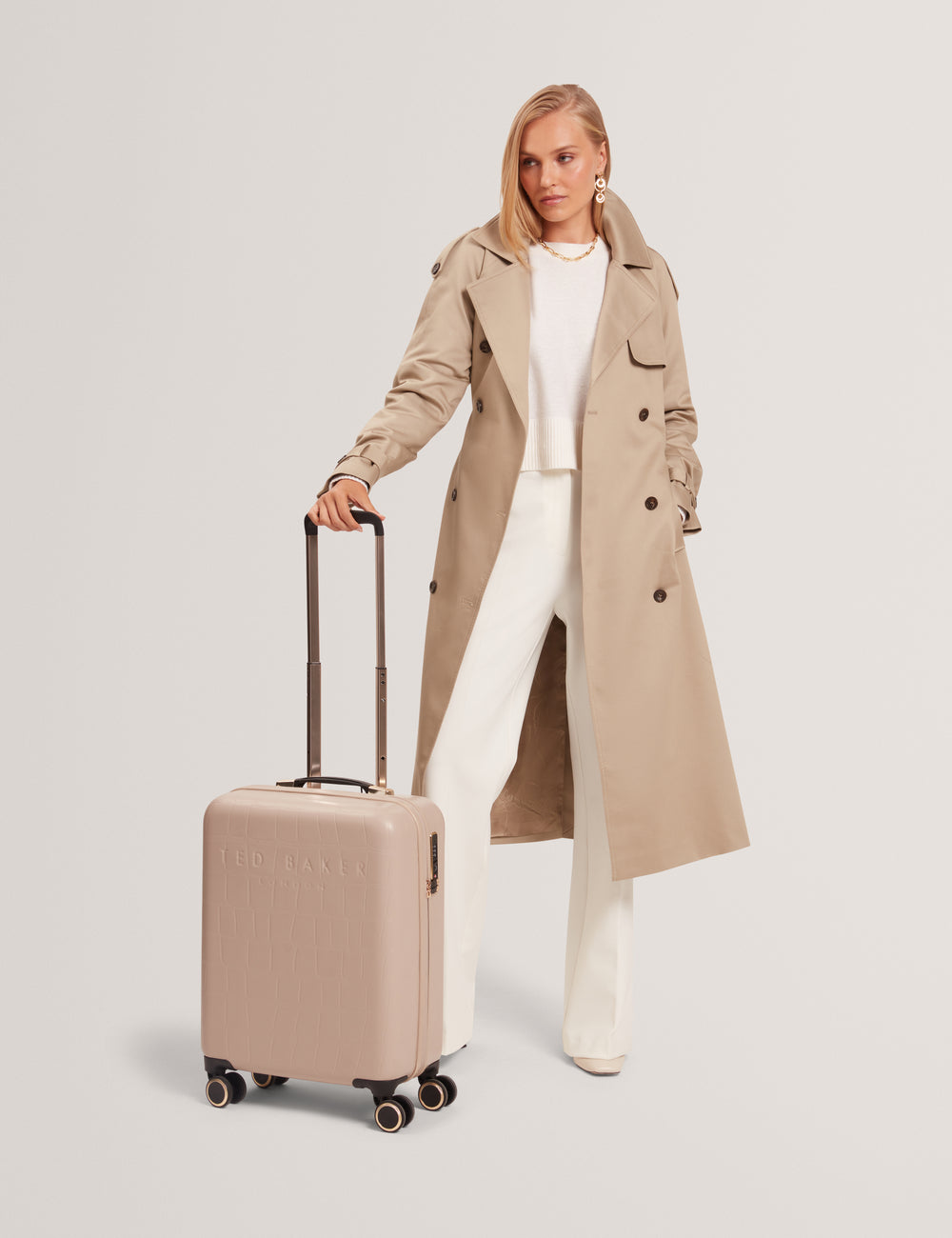 Model in a trench coat holding a beige suitcase on a light grey background