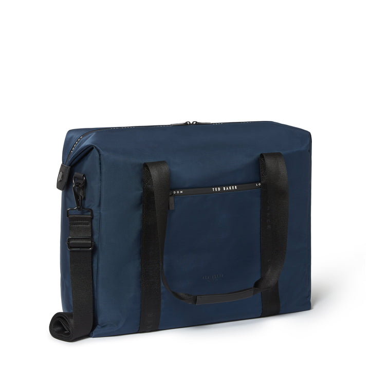 A navy blue duffel bag with black handles and a black adjustable shoulder strap, shown from a three-quarter angle on a white background. The brand name 'TED BAKER' is printed in white on a black horizontal strip on the front pocket.