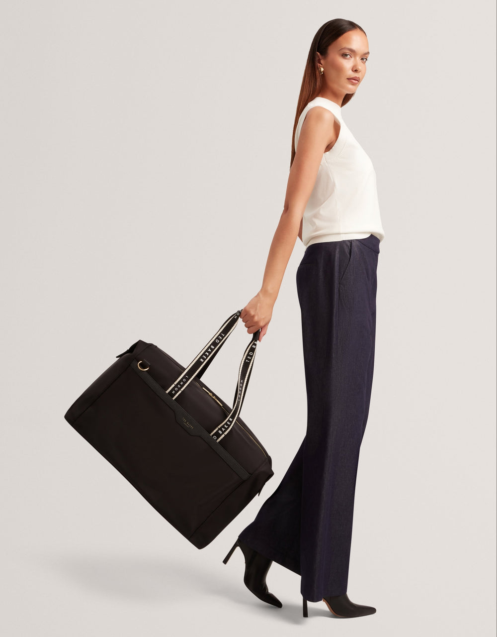 A woman in a white sleeveless top and dark navy wide-leg trousers carries a large black duffel bag with cream-tone handles against a light gray studio background.