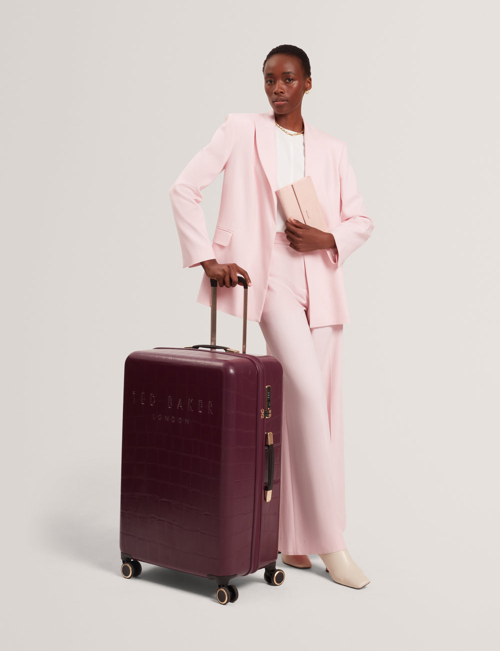 Model in a pink suit holding a plum coloured crocodile textured suitcase with 'Ted Baker London' branding on a light grey background