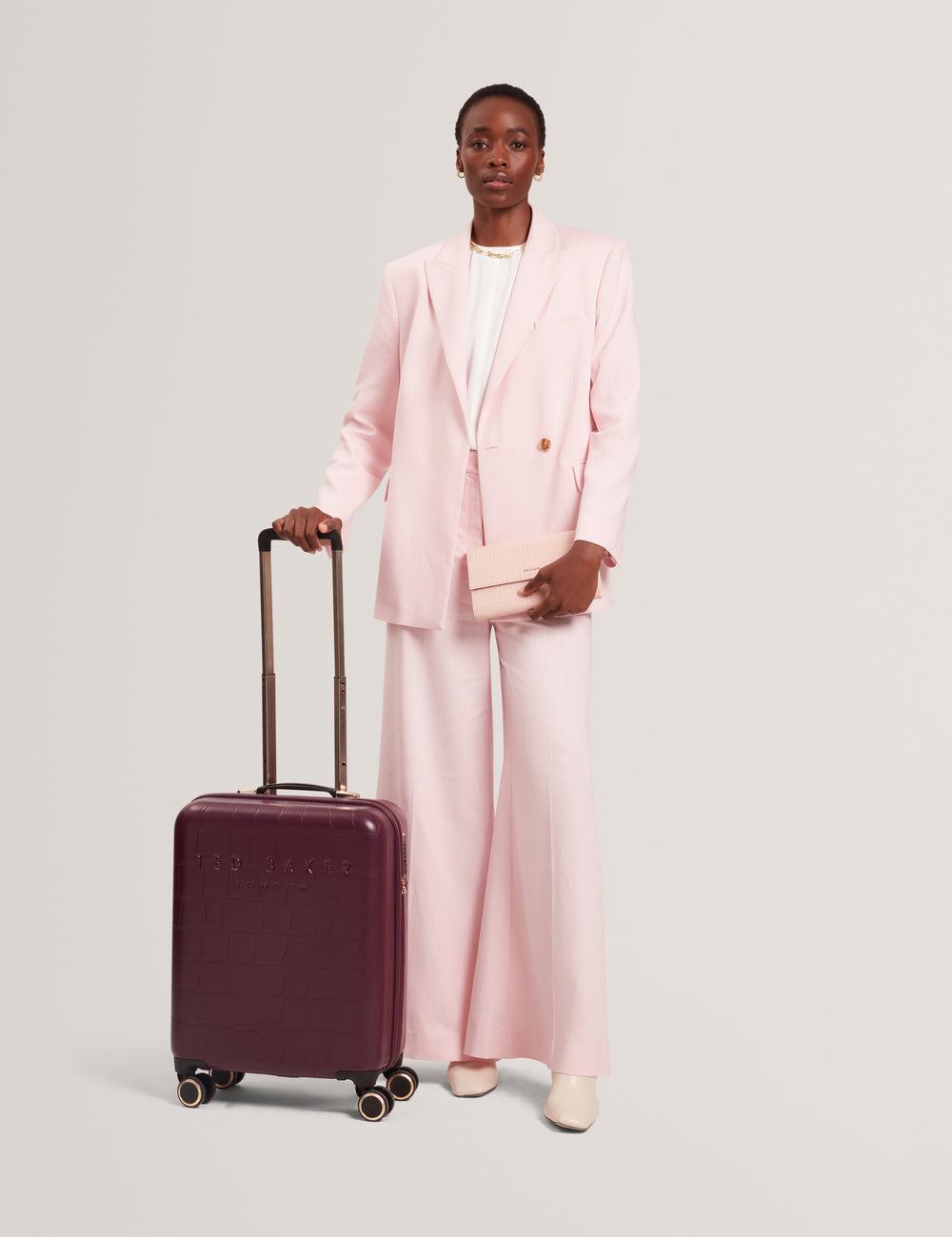 Model in a pink suit holding a plum colour suitcase on a light grey background