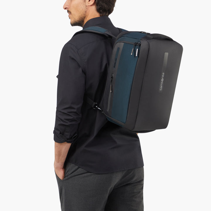 A man wearing a black long-sleeve shirt and gray trousers is shown from the side/back wearing a dark gray and navy backpack. The backpack has a vertical zippered compartment and a slim vertical logo on the side.